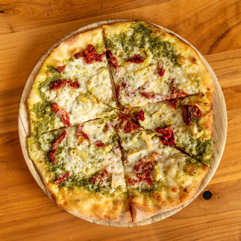 A sliced pesto pizza with sun-dried tomatoes, artichoke hearts, and melted mozzarella cheese on a wooden board.