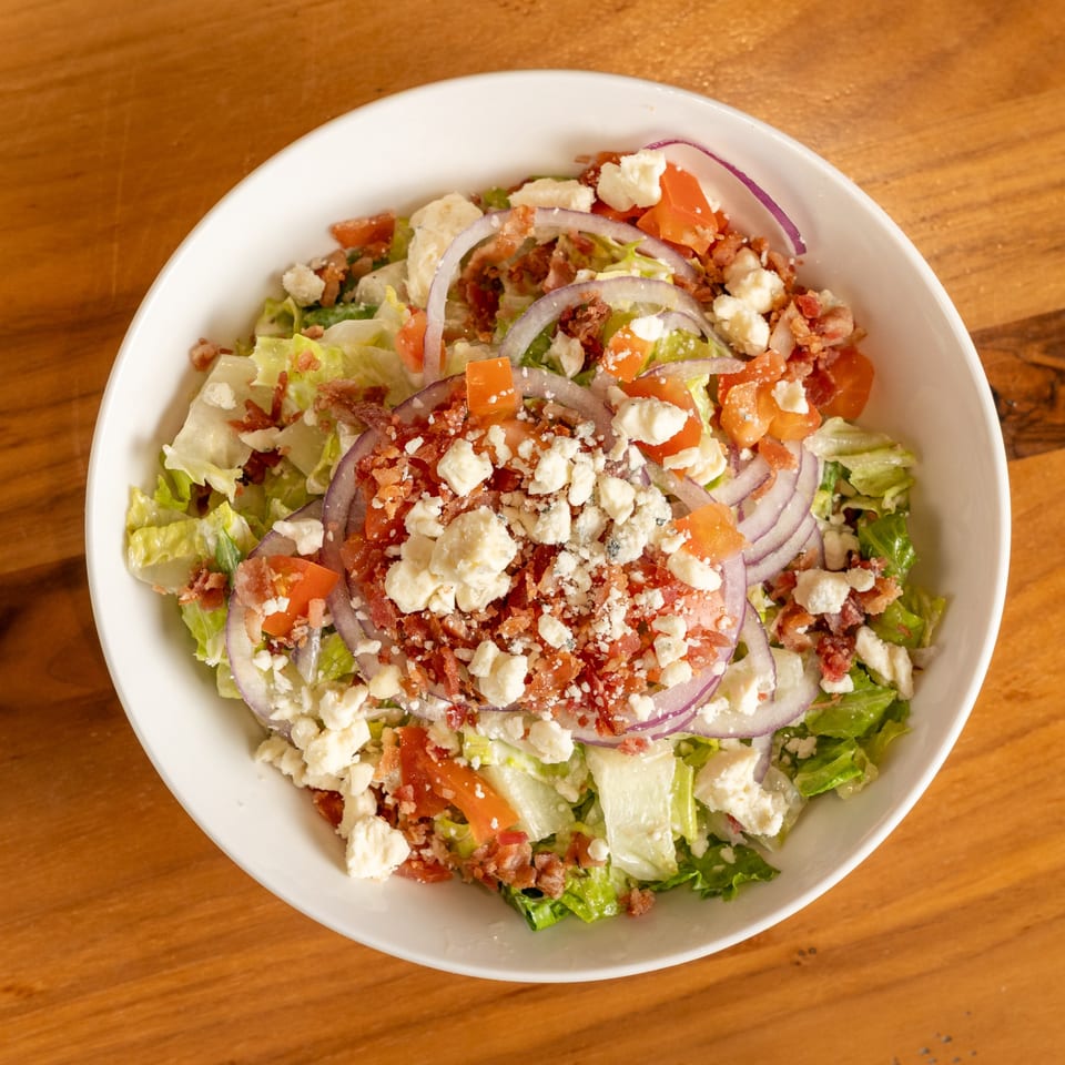 A white bowl filled with a fresh salad, topped with crumbled bacon, feta cheese, red onions, and tomatoes.