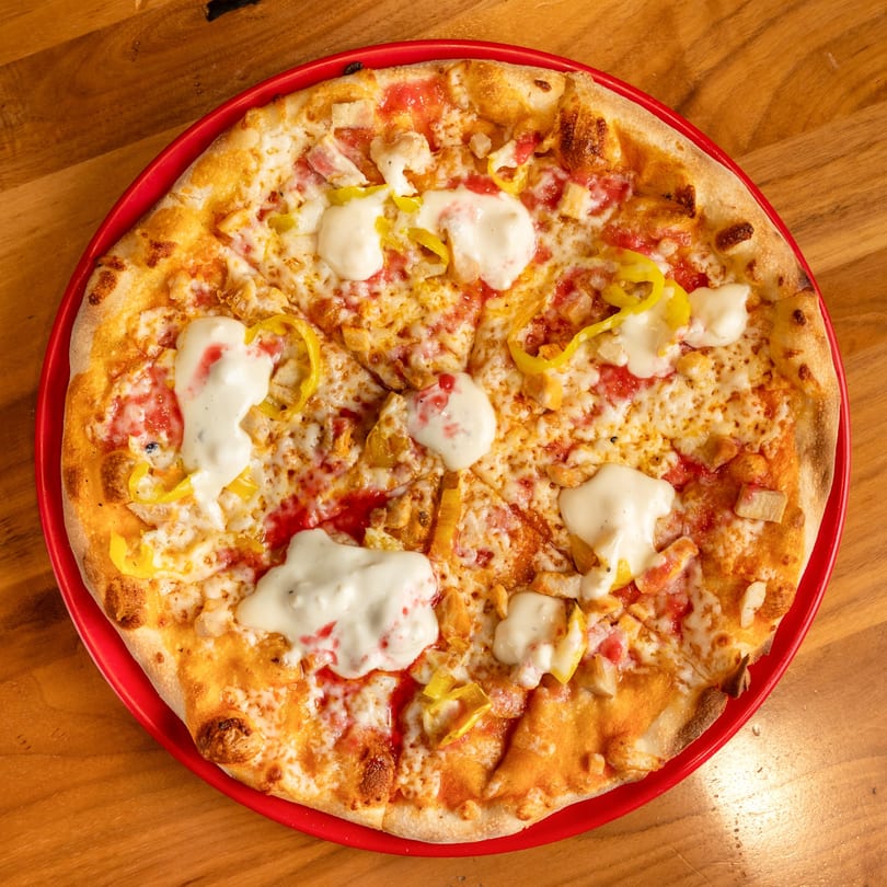 A whole pizza with chicken, yellow peppers, red sauce, cheese, and white sauce on a red plate.
