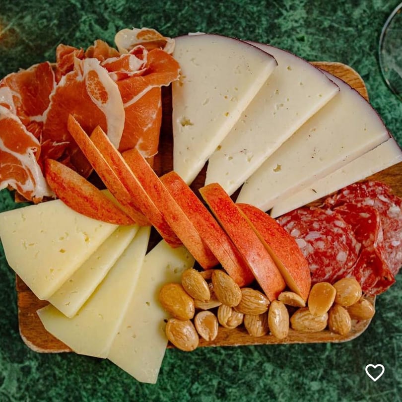 Charcuterie board with sliced cheeses, prosciutto, salami, apple slices, and almonds on a green surface.