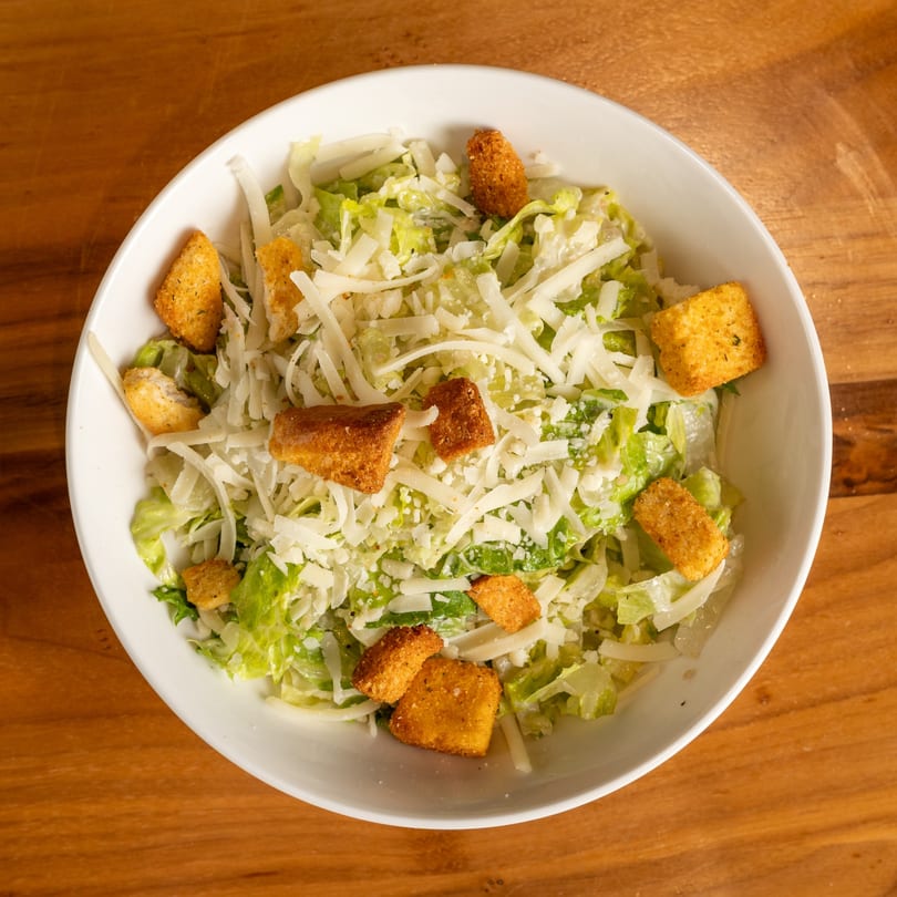 Caesar salad in a white bowl, topped with shredded cheese and croutons on a wooden table.